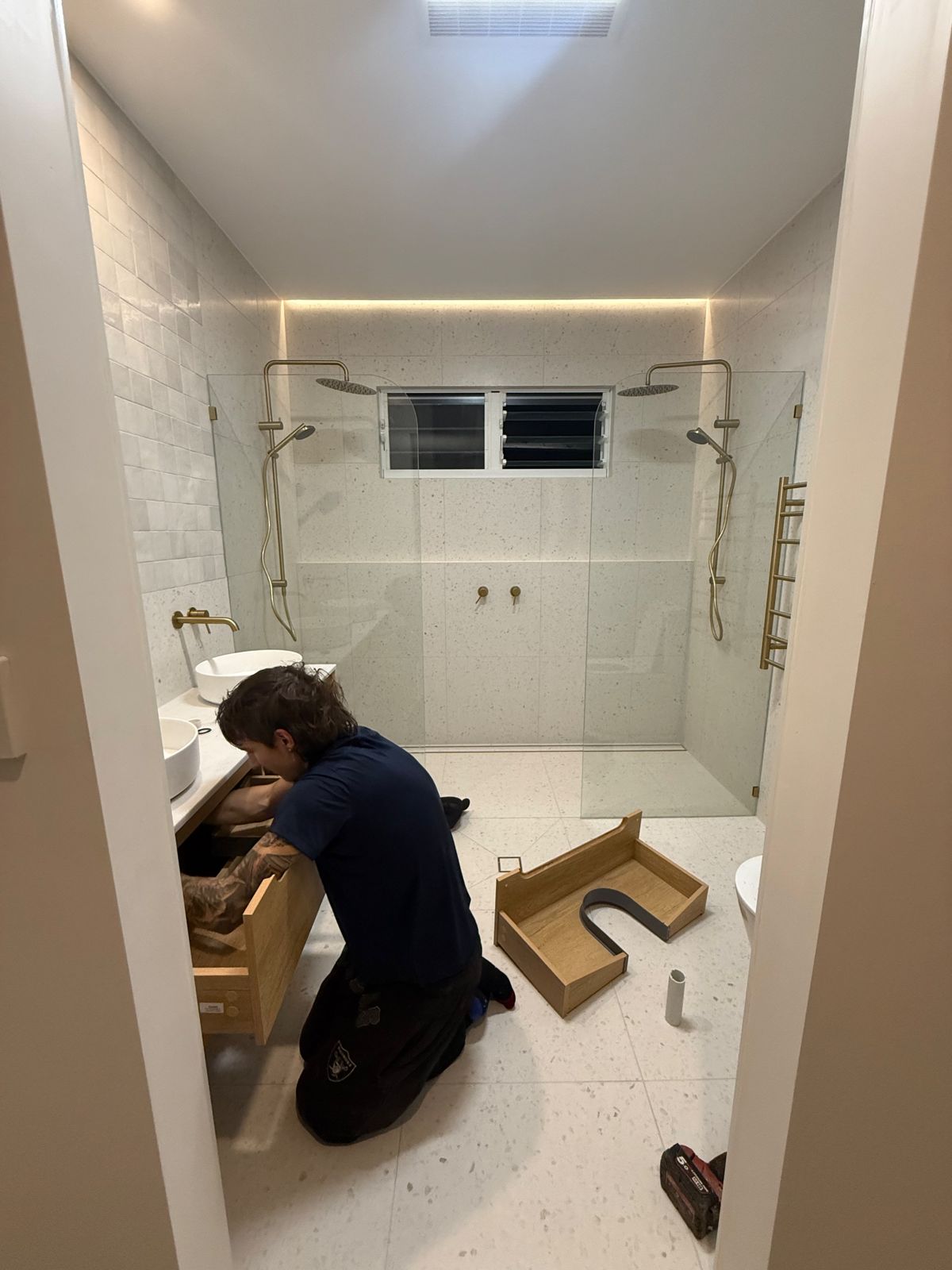 A tradesperson kneeling to fit the timber vanity in a newly tiled bathroom with a walk-in shower