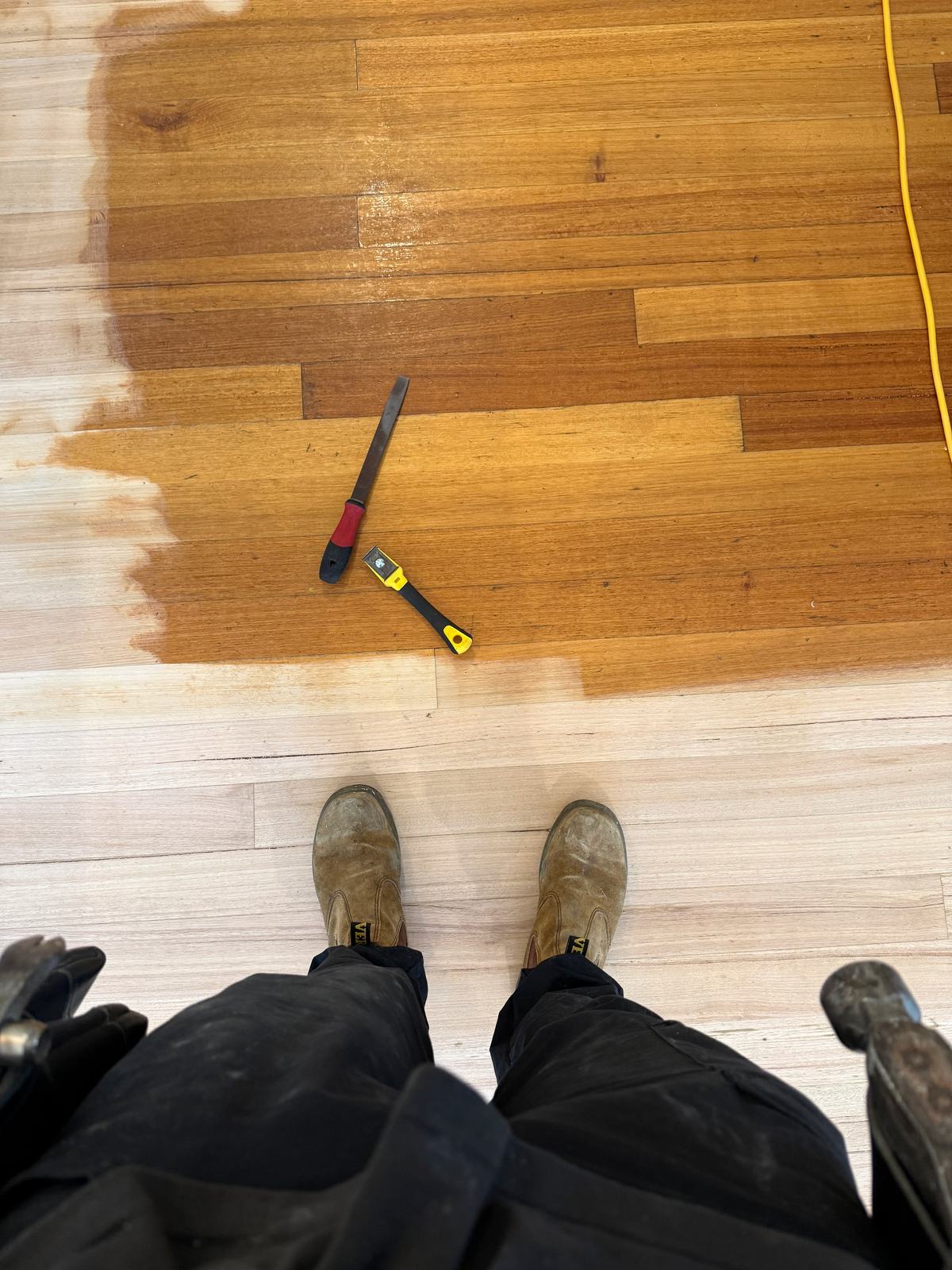 Looking down at work boots on freshly sanded hardwood, the first coat of finish turning the boards ahead a deep honey colour
