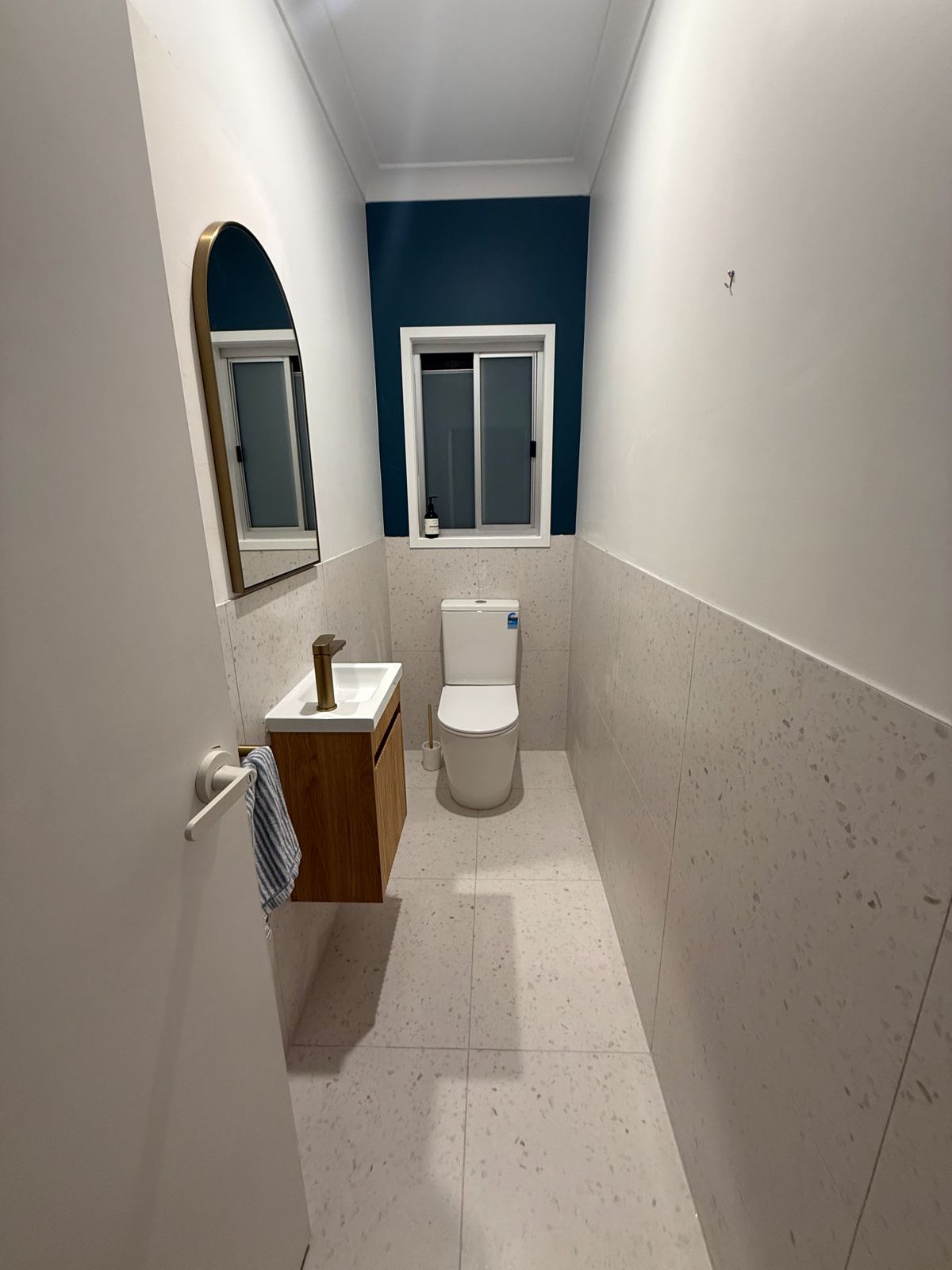 A narrow finished powder room with an arched mirror, small timber vanity and a dark feature wall
