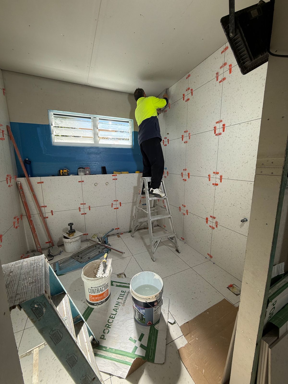 A tiler in hi-vis on a stepladder setting large pale wall tiles in a bathroom, levelling clips on every joint and a blue waterproofed wall beside him