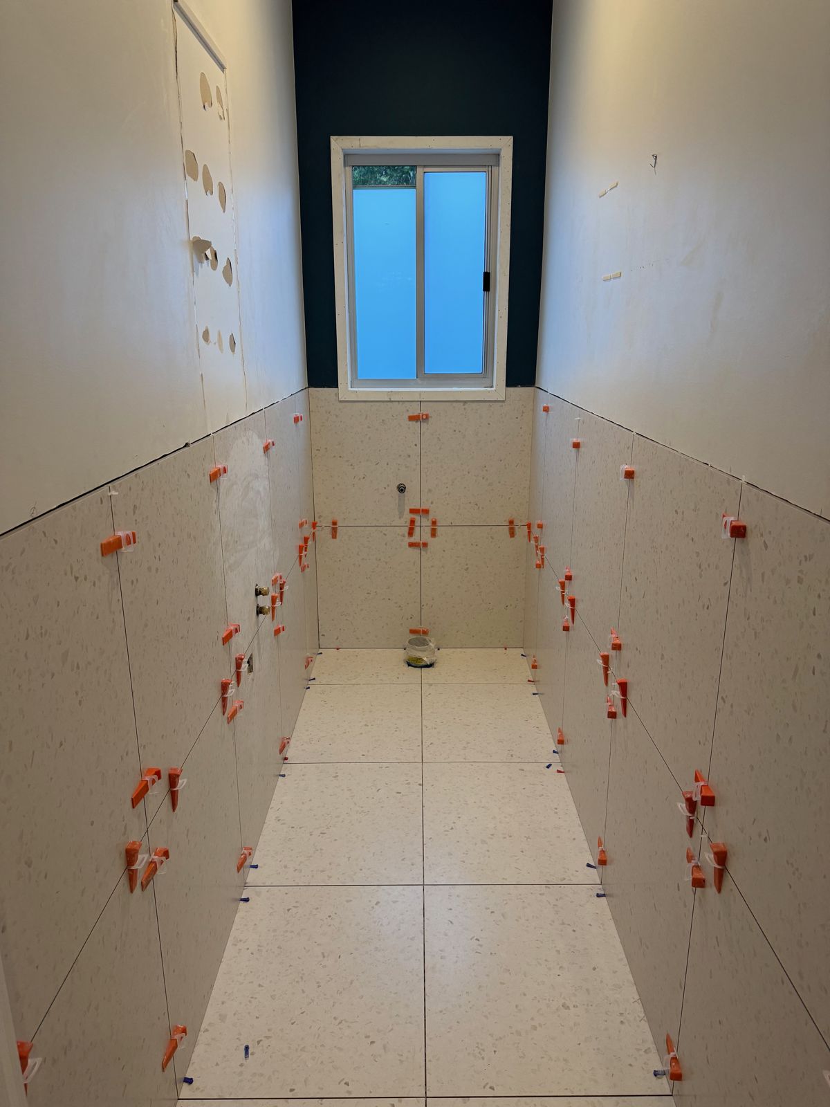 Large pale tiles going up in a long bathroom, red levelling clips at every joint
