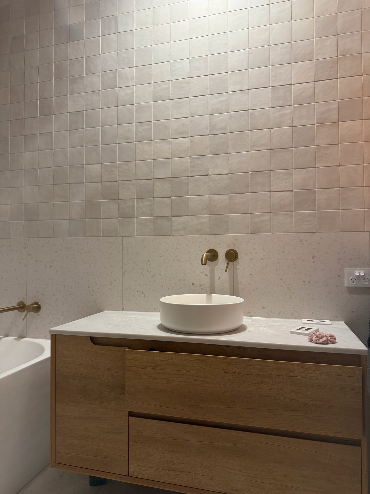 A timber vanity with a round white basin and brushed brass wall tapware under textured white wall tiles, the bath beside it