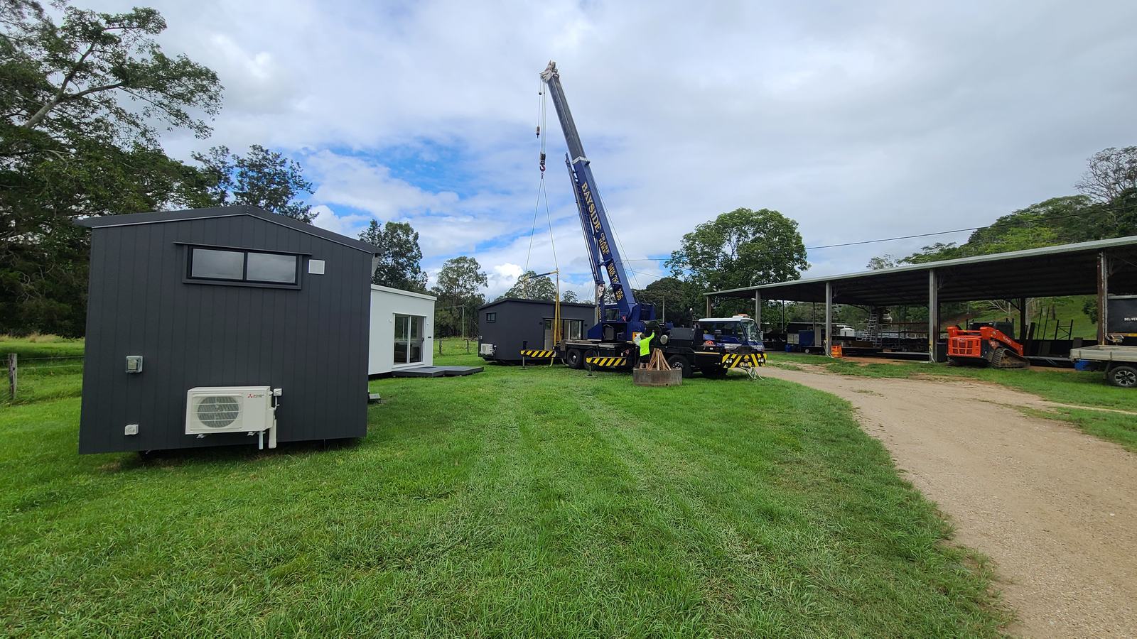 A blue mobile crane set up on a grassy yard beside three finished modular homes, a crew member in hi-vis at the outriggers