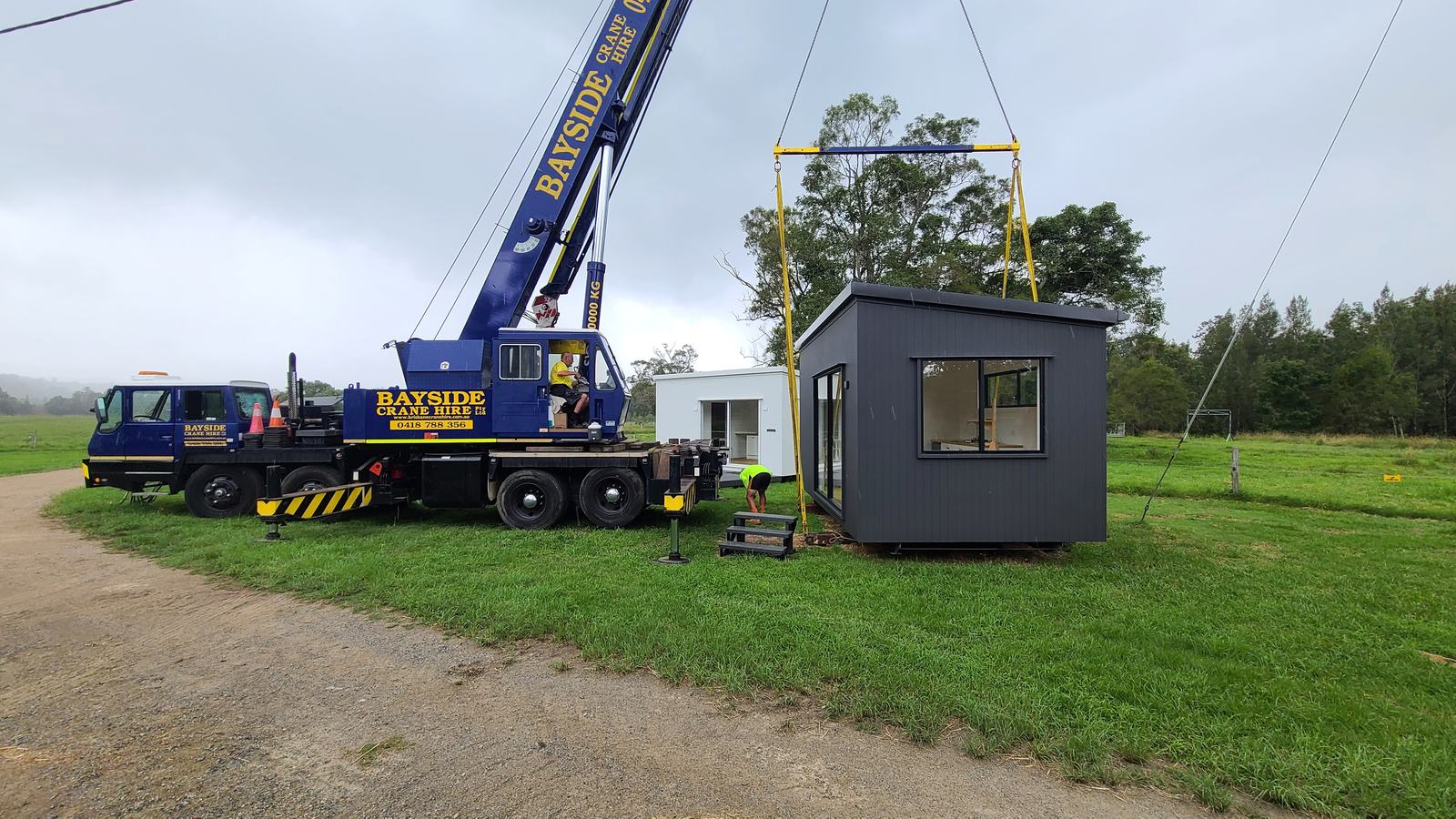 A mobile crane lifting a dark-clad modular home on a spreader bar while a crew member sets the stairs below