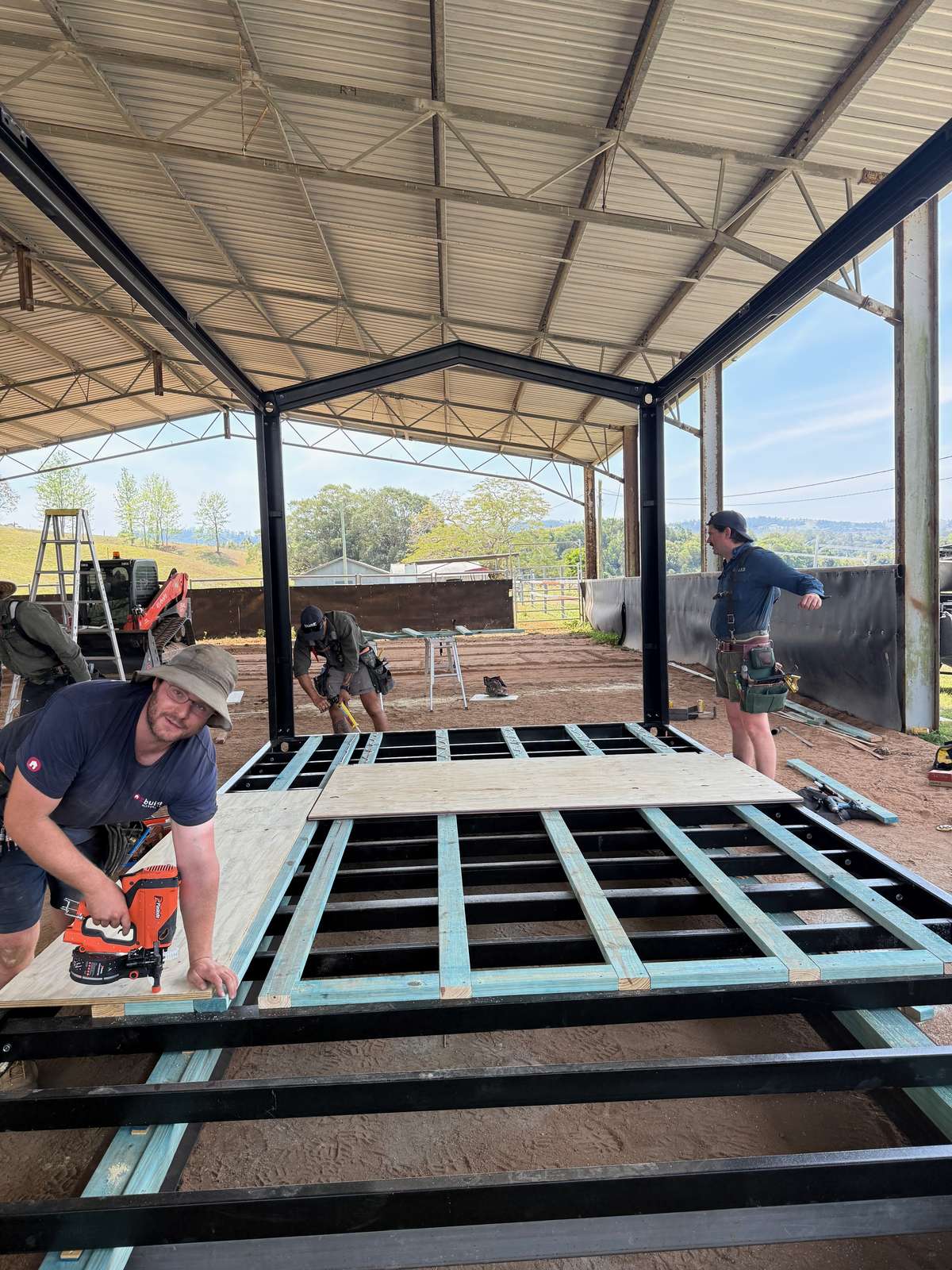 Builders fixing plywood flooring onto a steel floor frame under the shed, one with a nail gun