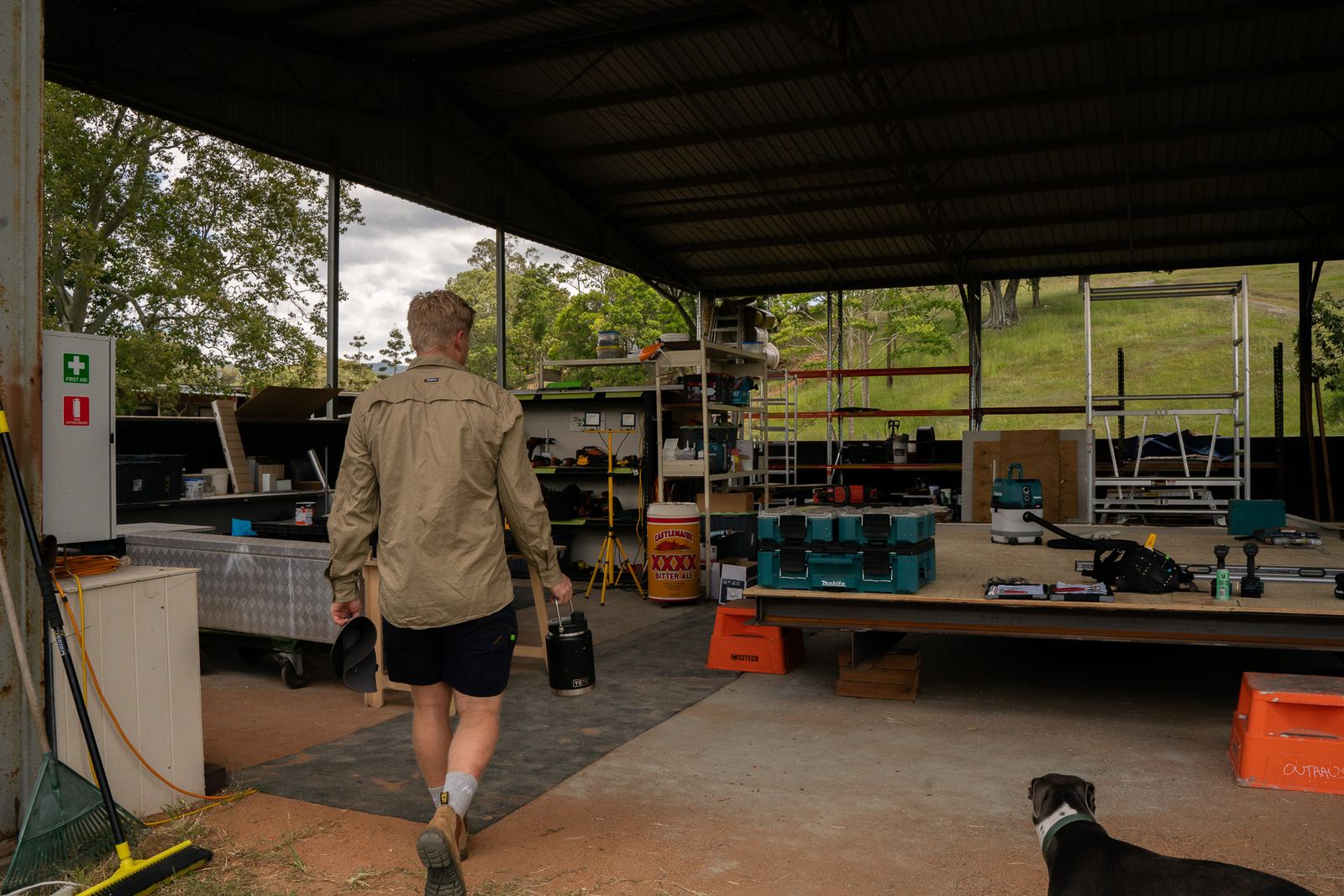 A builder in work gear walking into the shed past benches and scaffold, the site dog following