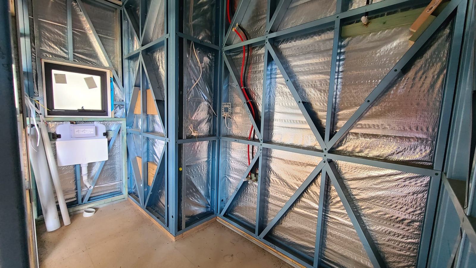 Inside an Outhaus module mid-build: the bathroom framed in blue light gauge steel with cross bracing, foil wrap behind and the window opening cut