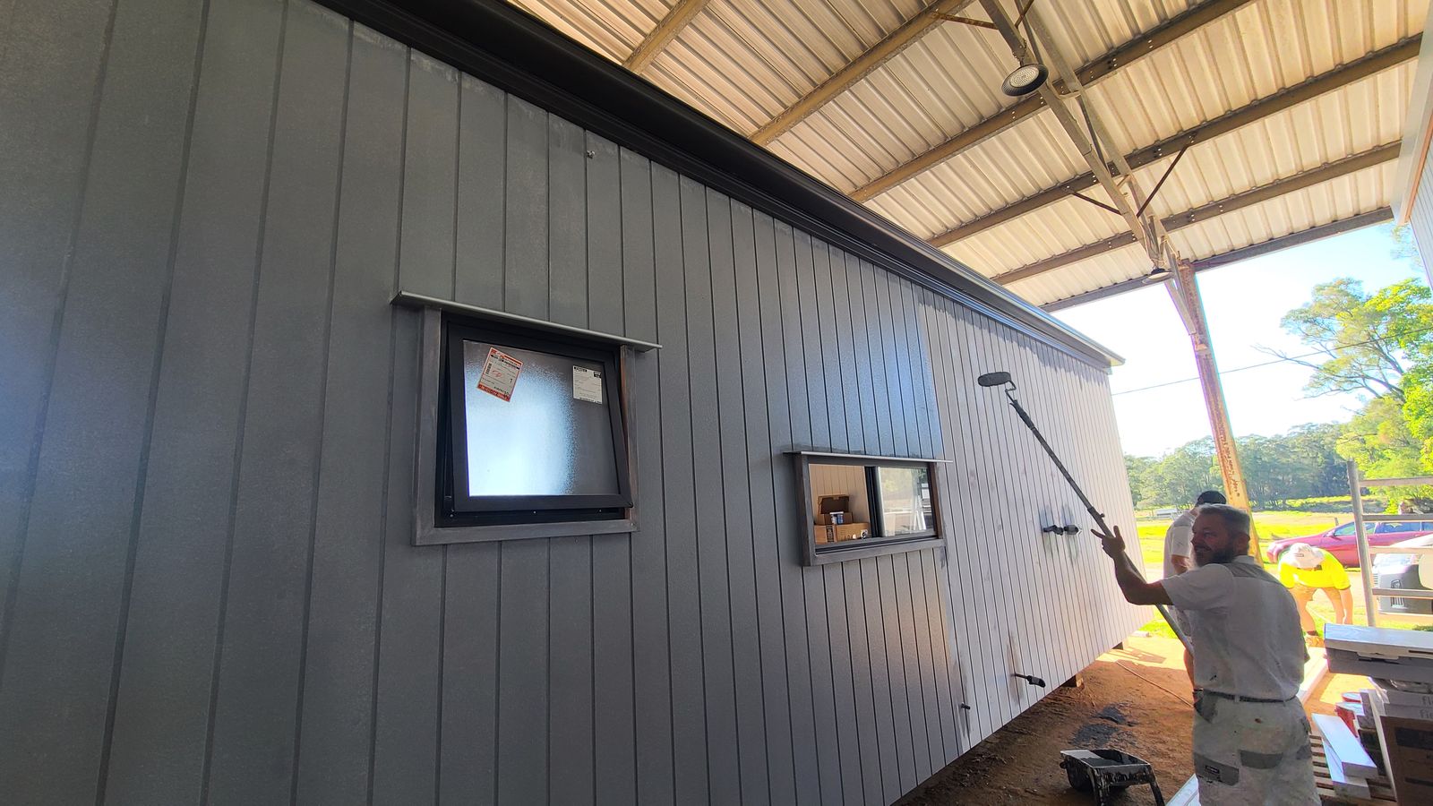 A painter rolling a fire-rated coating onto the dark cladding of a modular home in the factory shed