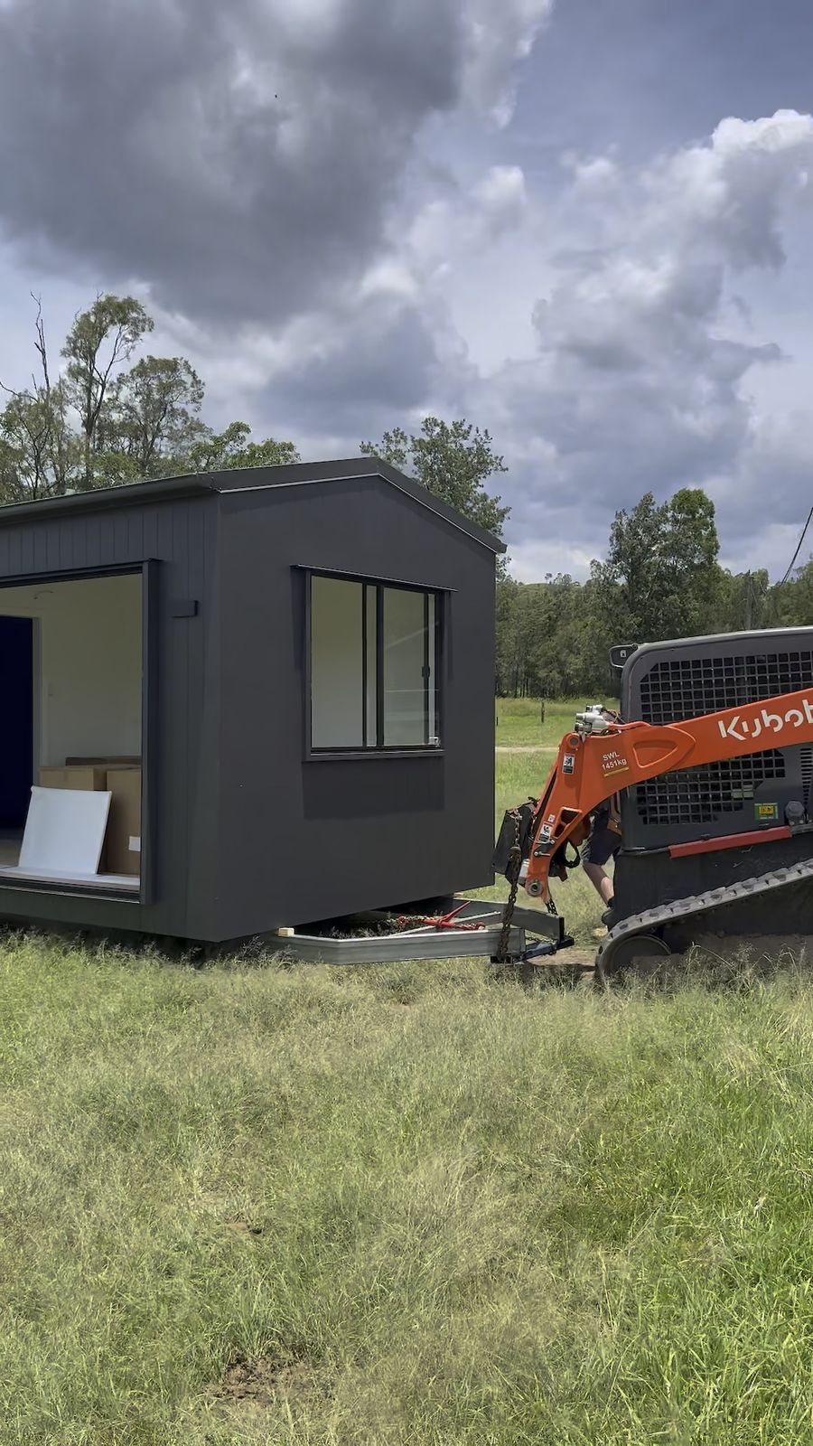 A tracked skid steer easing a dark-clad module into position on the grass