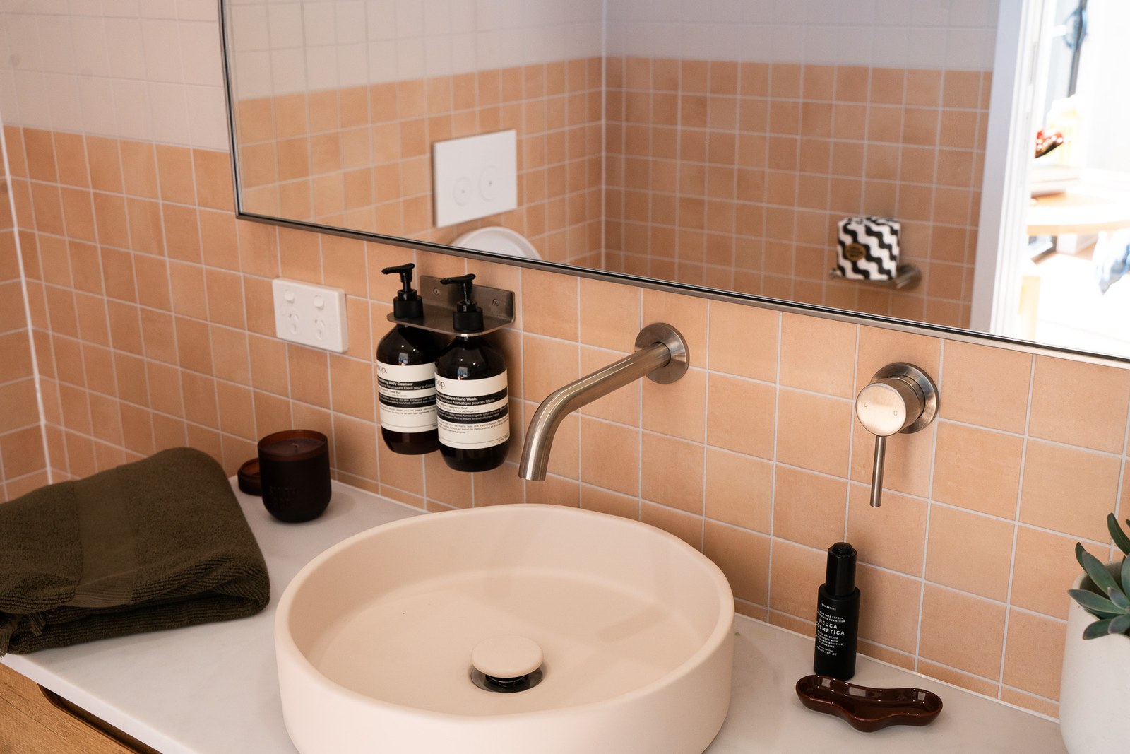 A round stone basin on a vanity with a wall-mounted tap, pink square tiles and a large mirror