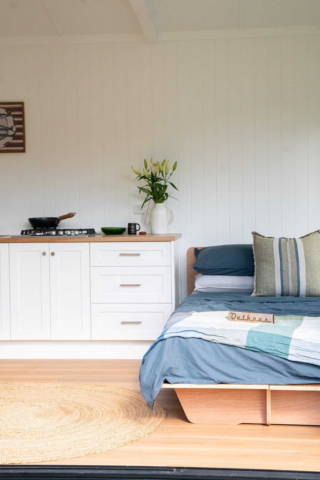 Inside a white second home, a made bed beside a white shaker kitchen with a timber benchtop and a jar of lilies