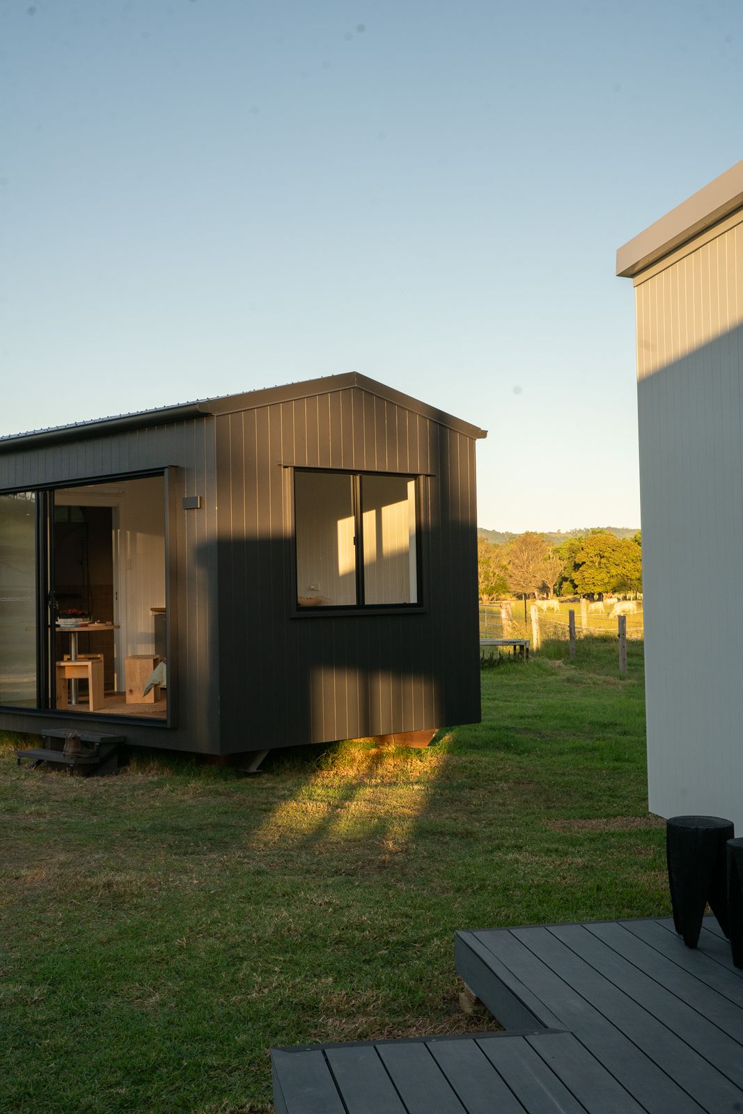 A dark-clad modular second home on a grassy block in late afternoon sun, with a step down to a deck
