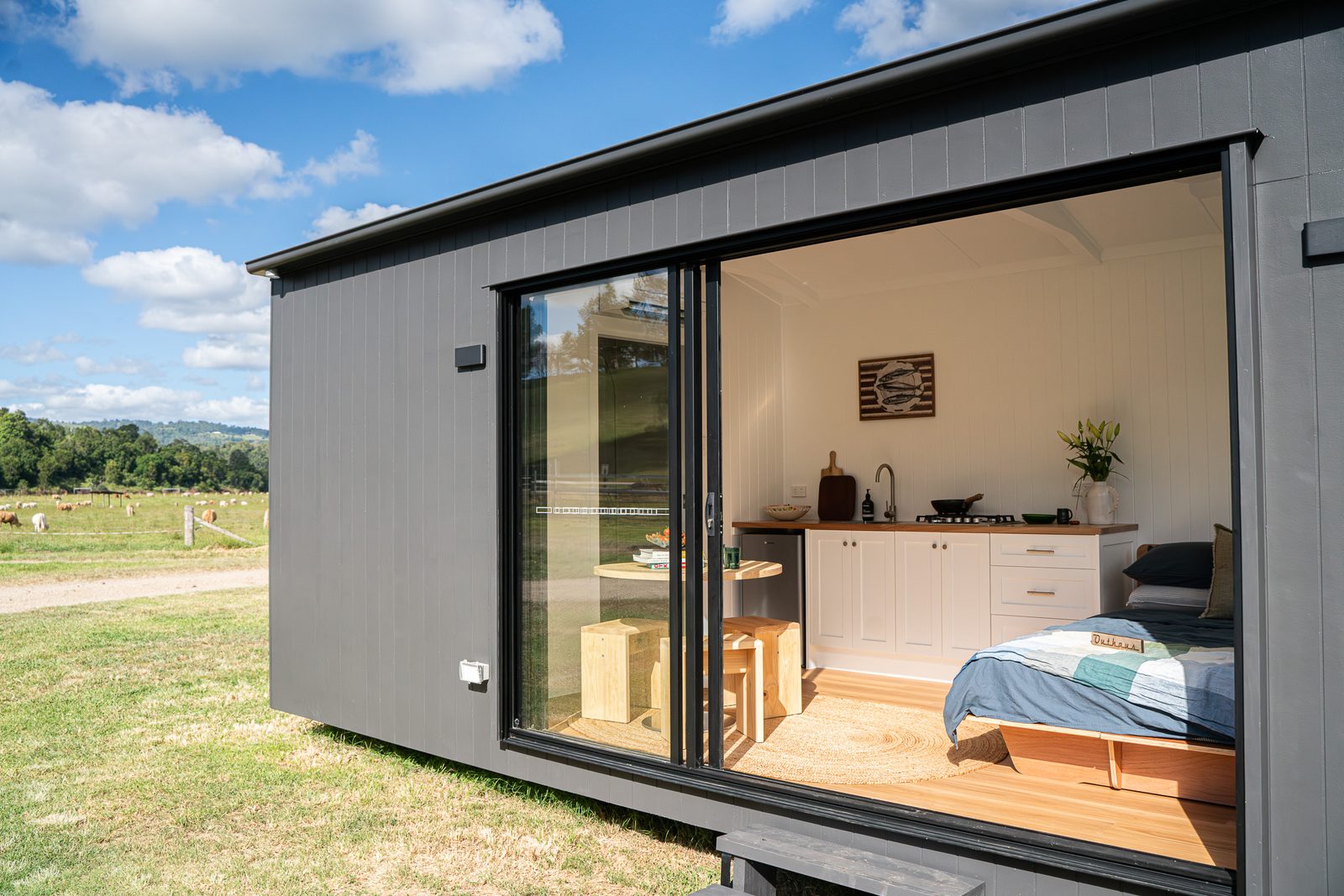 A dark-clad second home with its glass slider open, showing a made bed and kitchen inside, on a grassy paddock