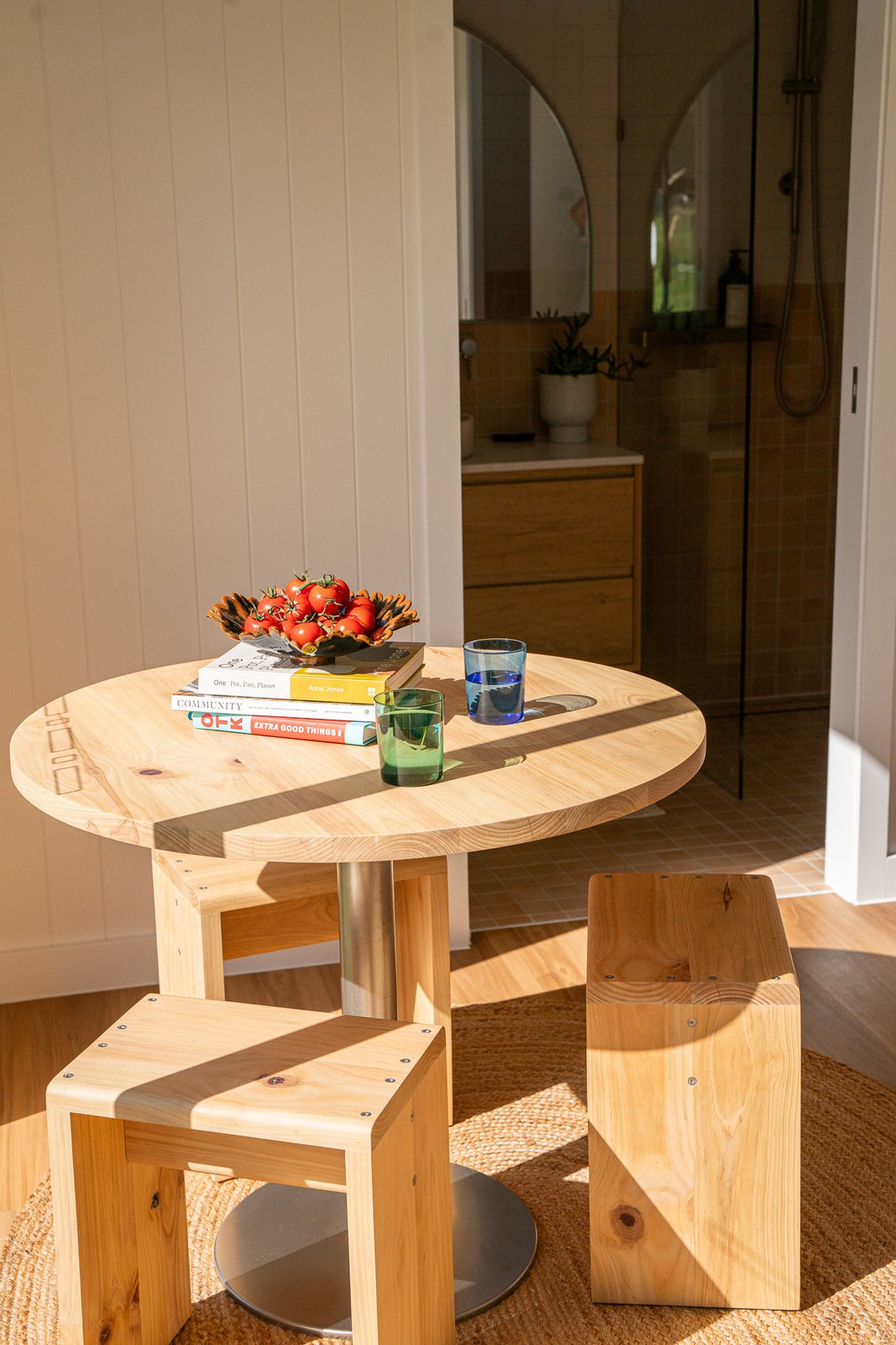 A round timber table with two timber stools, a bowl of fruit and books, the bathroom visible through the doorway behind
