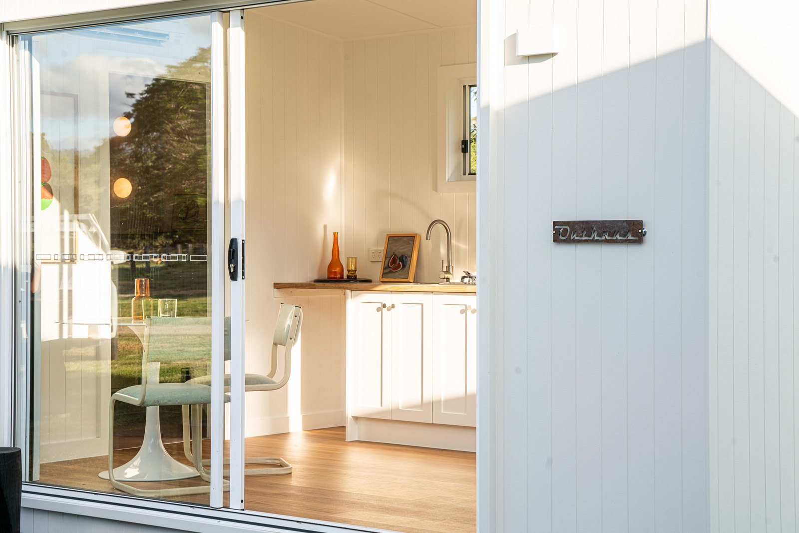 Looking through open glass sliding doors from the deck into a white second home, a round table and the kitchen inside