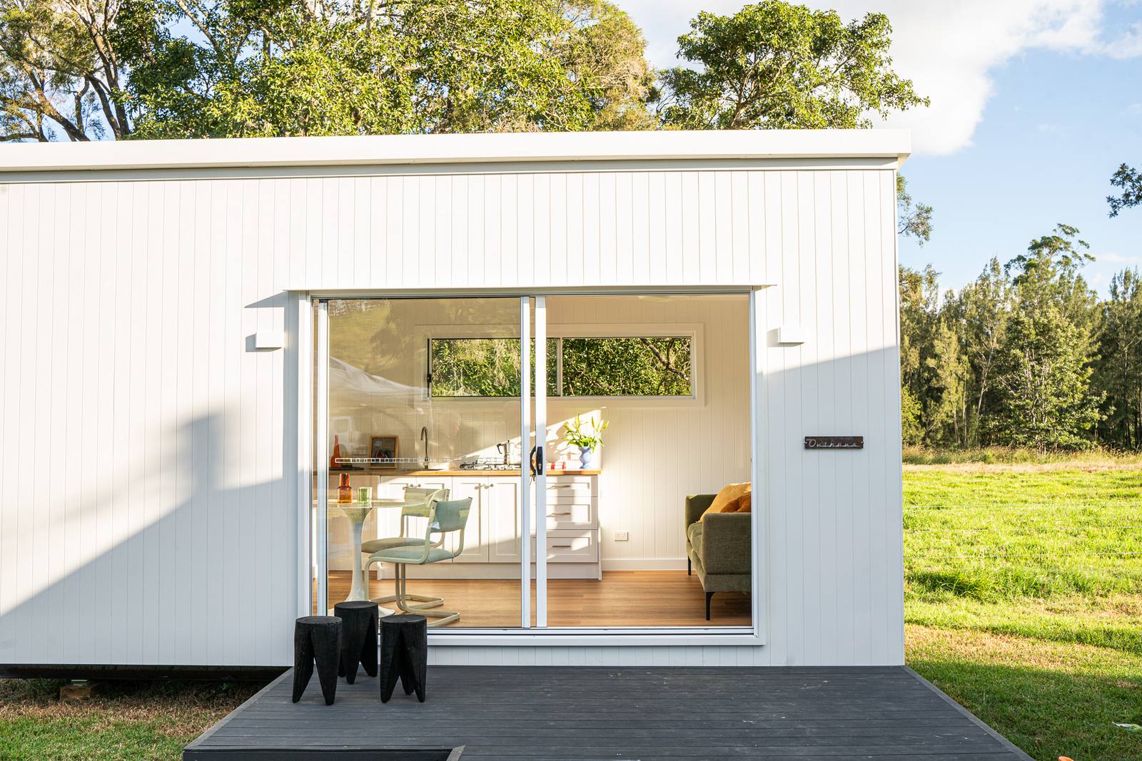 A white-clad modular second home with its sliding doors open onto a dark timber deck, on a green paddock