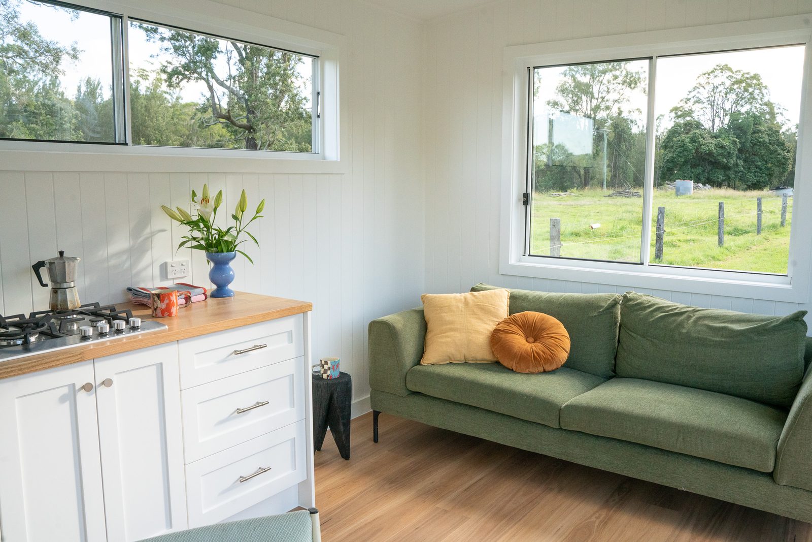Inside a white second home, a green sofa under two windows beside a white kitchen with a timber benchtop