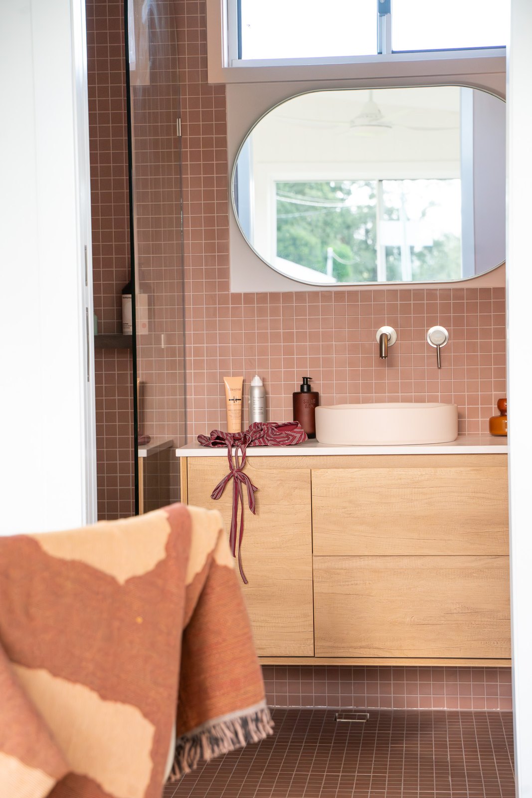 A bathroom with deep pink square tiles, an oval mirror, a timber vanity and a round basin with a wall tap