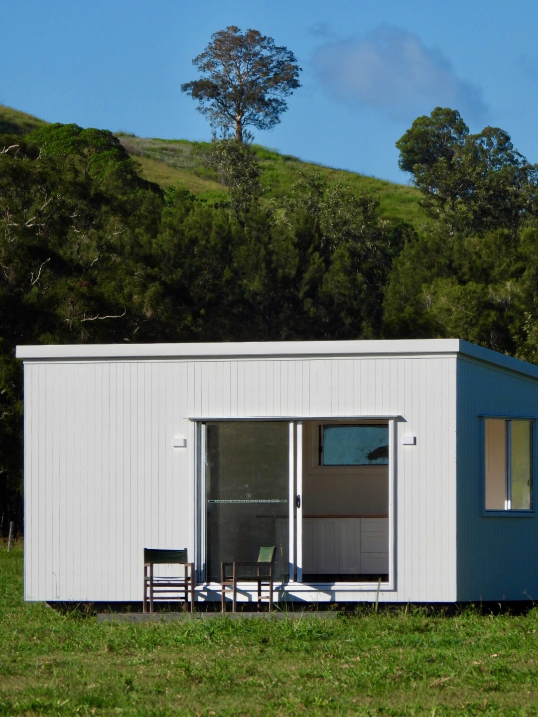 A white-clad modular second home with sliding doors and two chairs out front, under a green hill and blue sky