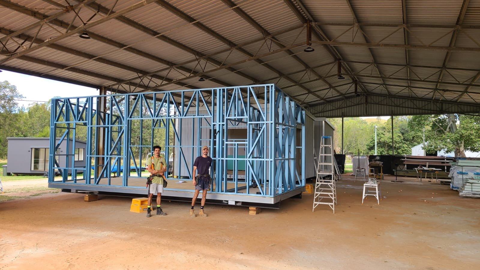 Two builders standing in front of a blue steel-framed module under a big open shed
