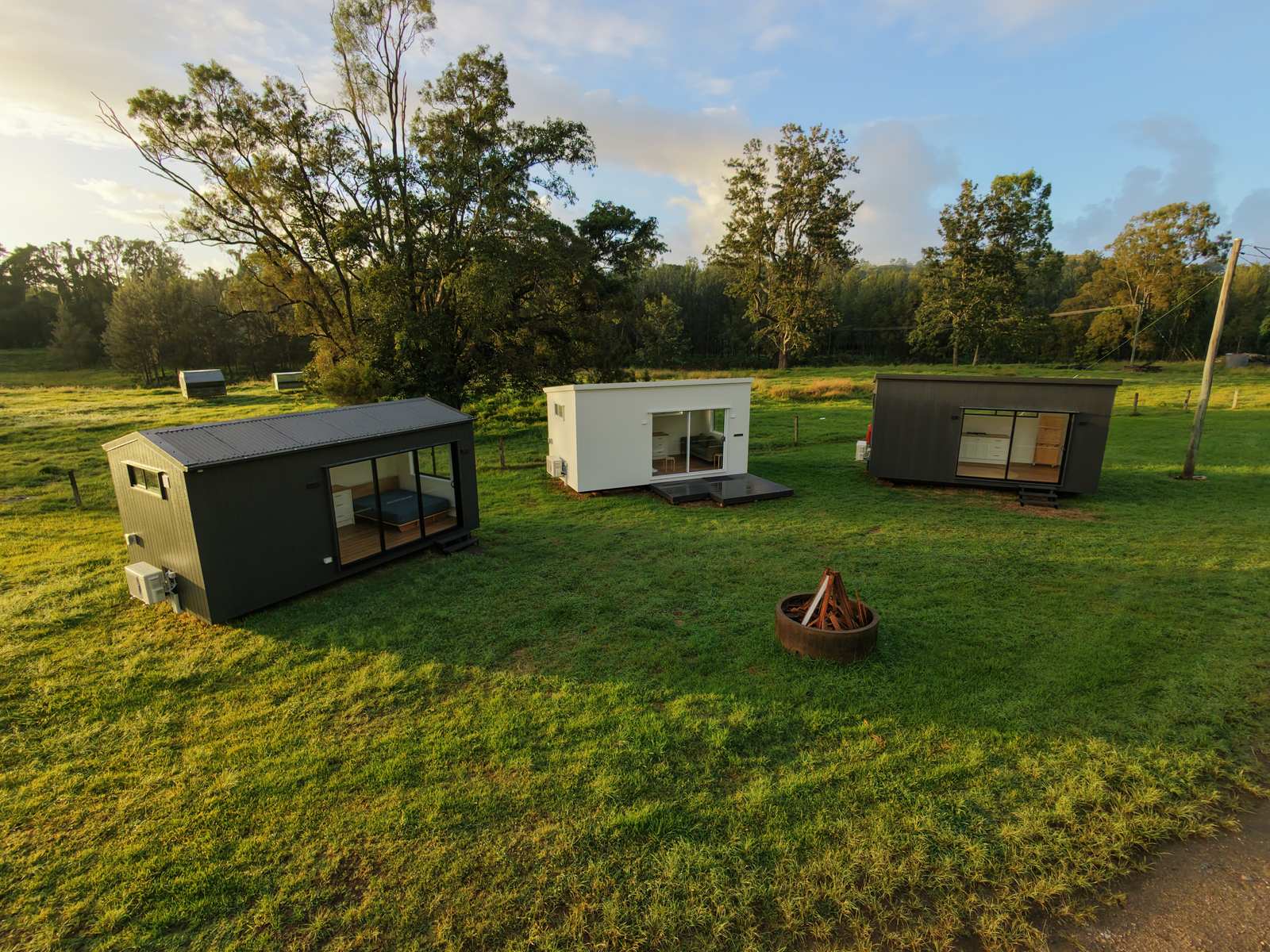 Drone view at sunrise of three finished modular second homes, two dark and one white, around a fire pit on green grass